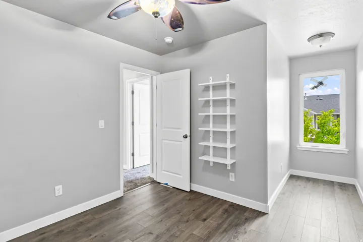 Interior room featuring wood-finish flooring, a ceiling fan with integrated lighting, a built-in shelving unit, and a window providing natural light