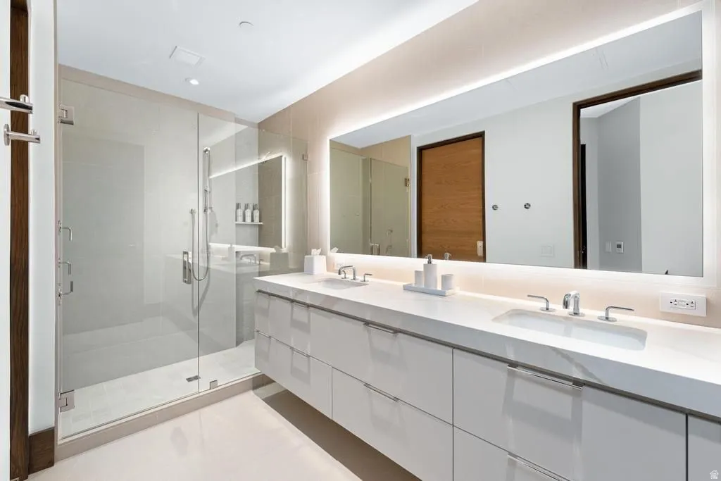Contemporary bathroom featuring a spacious glass-enclosed shower with a handheld fixture, an expansive double vanity with integrated sinks, white cabinetry, and a large illuminated mirror