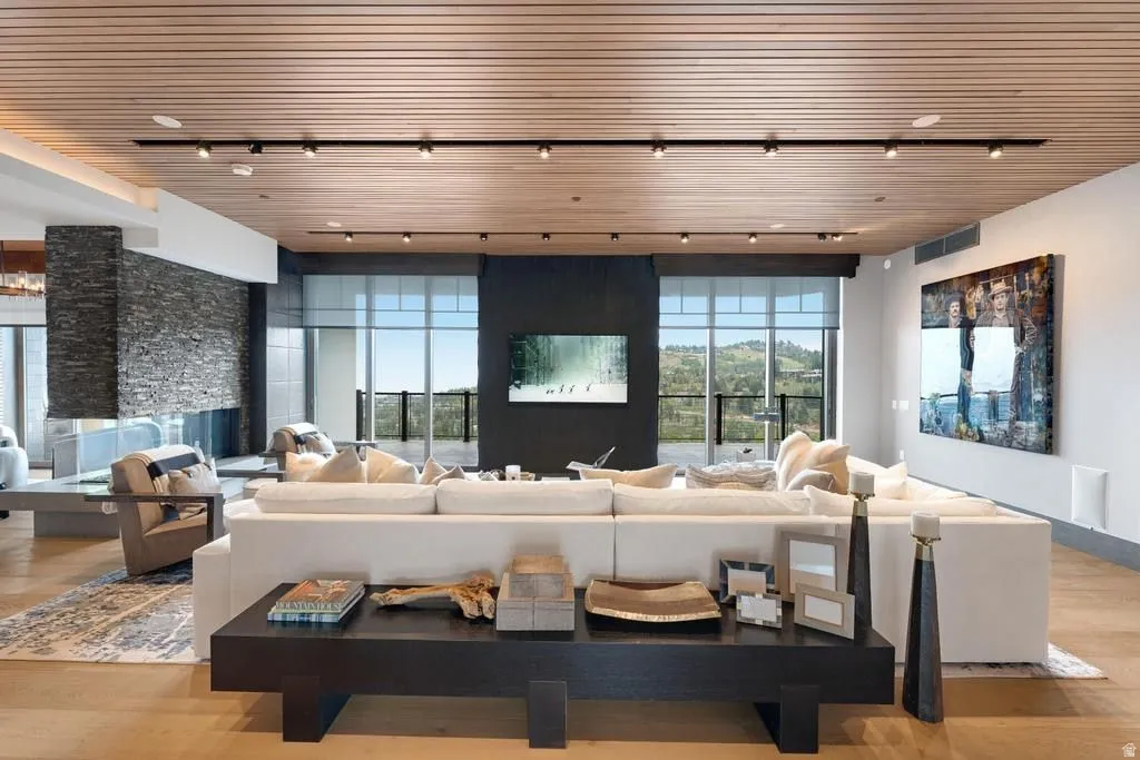 Spacious living area featuring a stone-clad fireplace, wood-finish ceiling with track lighting, multiple large windows, and light wood-finish flooring