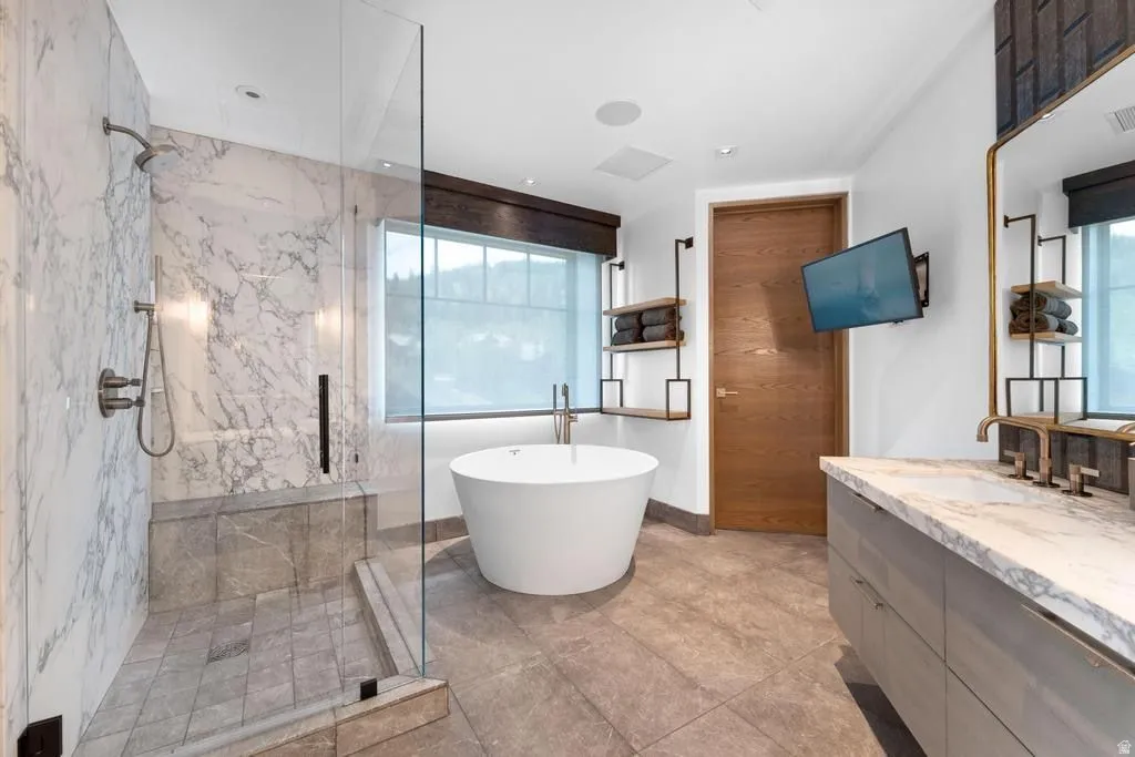 Spacious bathroom featuring a freestanding soaking tub, a walk-in shower with clear glass enclosure and marble-look wall tiling, a double vanity with a stone countertop, and wood-finish flooring