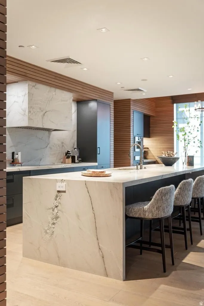 Gourmet kitchen featuring a waterfall island with integrated power, custom dark cabinetry, wood-finish wall paneling, full-height stone backsplash, and recessed lighting