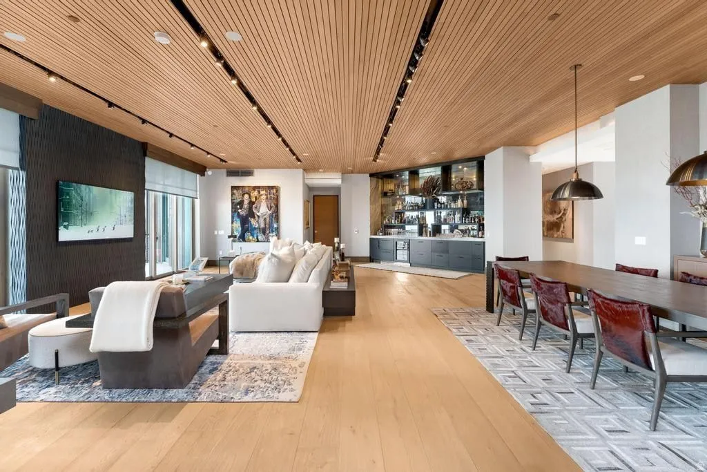 Open-concept living space featuring wood-finish flooring, a slatted wood-panel ceiling with integrated track lighting, a built-in wet bar with dark cabinetry, and a formal dining area with a rectangular table