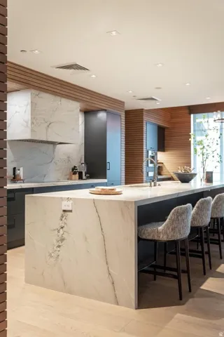 Gourmet kitchen featuring a waterfall island with integrated power, custom dark cabinetry, wood-finish wall paneling, full-height stone backsplash, and recessed lighting