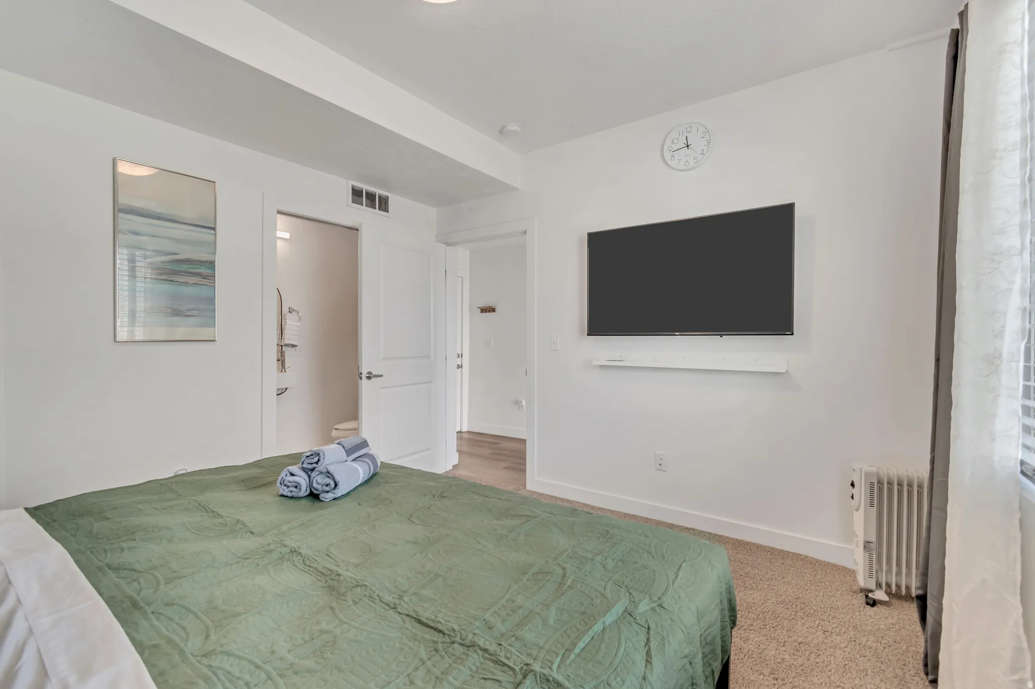Carpeted bedroom featuring a wall-mounted television, a wall-mounted clock, and a window with blinds and sheer curtains