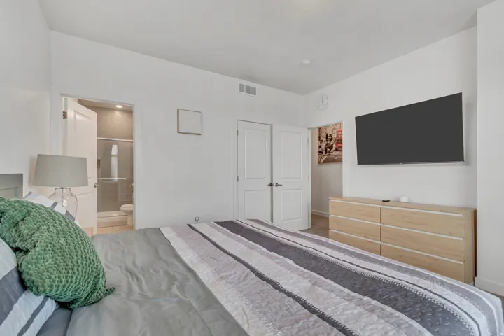 Bedroom interior featuring a neutral color palette, carpet flooring, a wall-mounted entertainment area, and an en-suite bathroom with a glass-enclosed shower