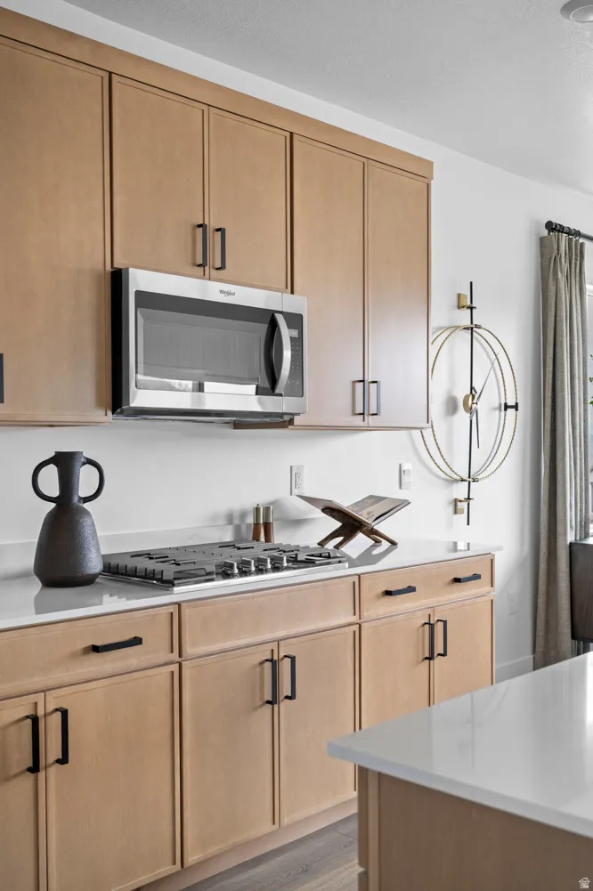 Contemporary kitchen featuring light wood-finish cabinetry with black hardware, stainless steel microwave, gas cooktop, and light-toned countertops