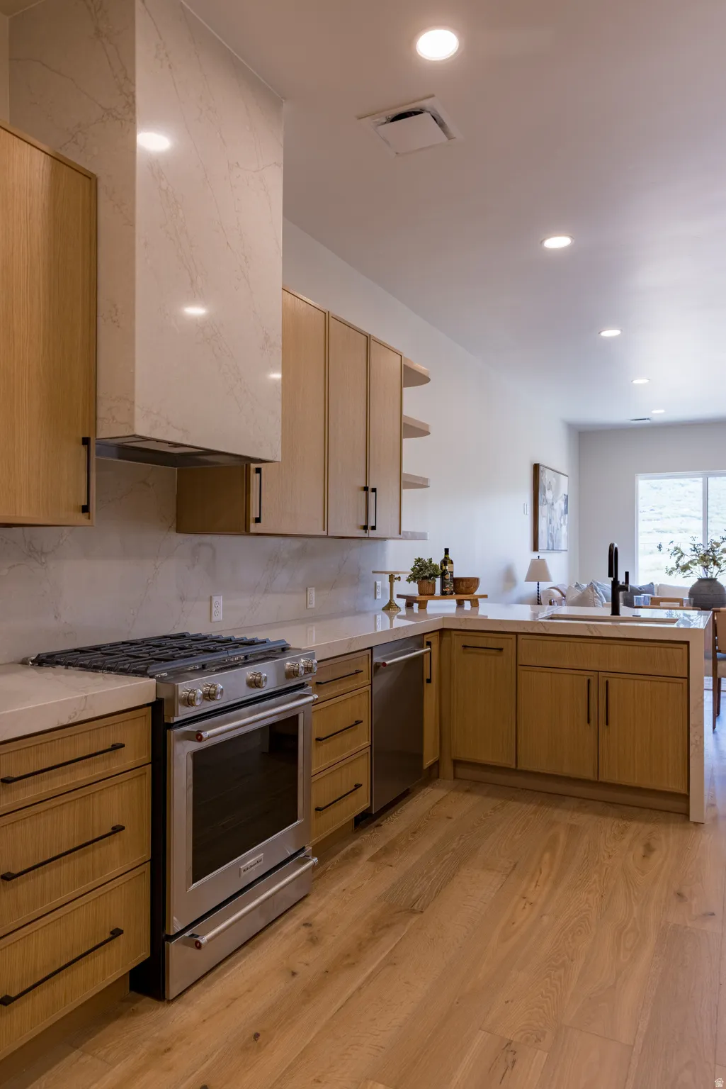 Modern kitchen featuring light wood-finish cabinetry with black hardware, stainless steel appliances, a stone countertop, and a matching stone backsplash and range hood