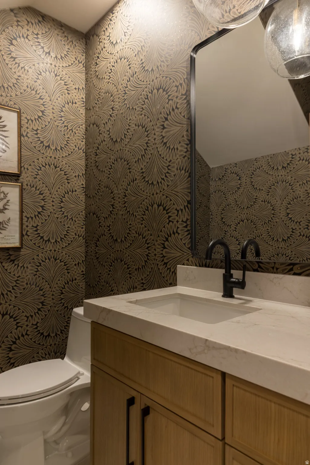Powder room featuring intricate patterned wallpaper, a light wood-finish vanity with dark hardware, a white countertop with an integrated sink, a matte black faucet, and an arched mirror
