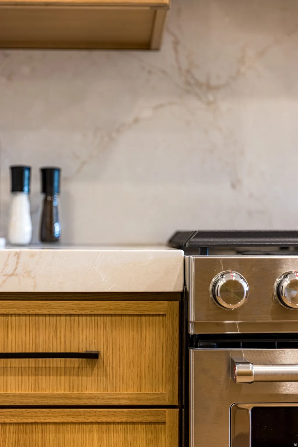 Integrated stainless steel range with metal knobs, light wood cabinetry featuring black hardware, a light-toned countertop, and a coordinating backsplash with subtle veining