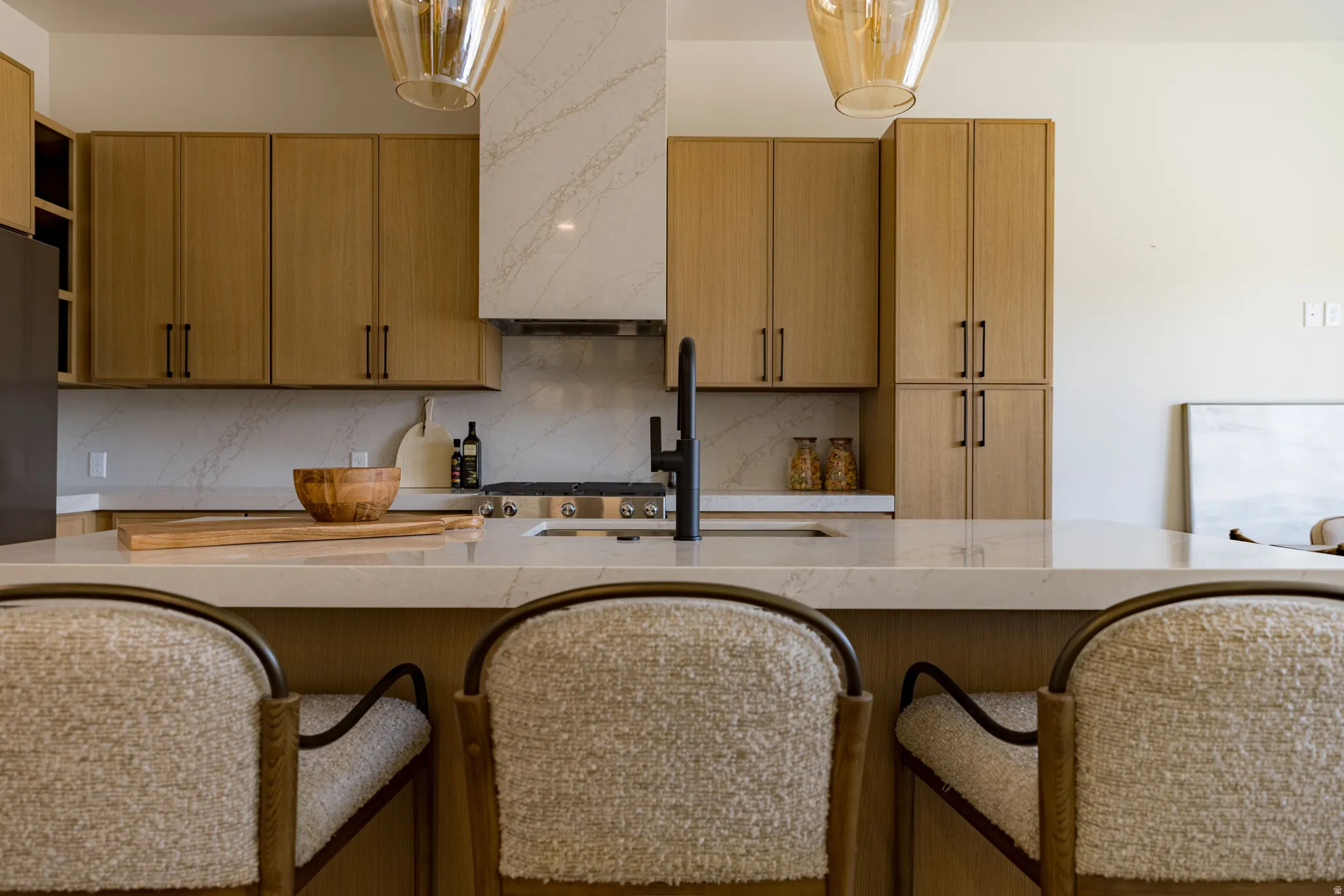 Modern kitchen featuring light wood-finish cabinetry, a large kitchen island with integrated sink, quartz countertops, a matching quartz backsplash, and a built-in range with an integrated hood