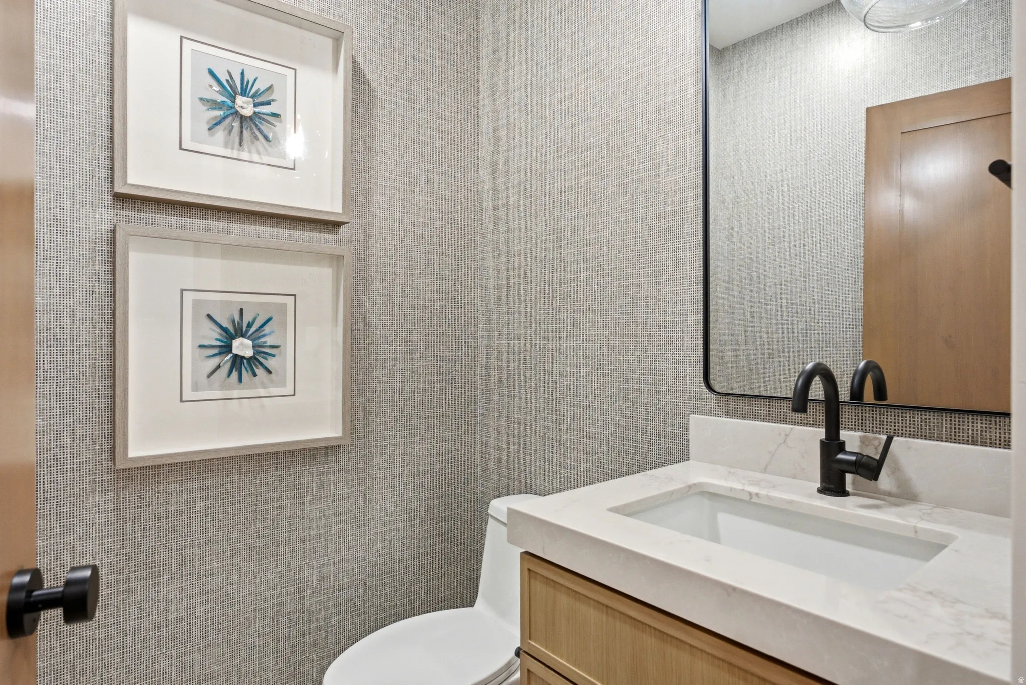 Powder room featuring textured wall covering, a vanity with a light wood finish, a white countertop, an undermount sink, and a matte black faucet