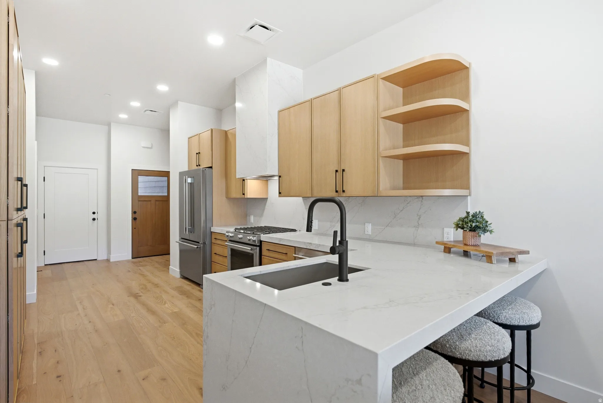 Modern kitchen featuring a waterfall-edge island with integrated sink and matte black faucet, stainless steel appliances, light wood-finish cabinetry, built-in shelving, and light wood-finish flooring