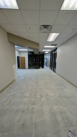 Spacious interior corridor featuring suspended ceiling tiles with integrated fluorescent lighting, exposed concrete flooring, and multiple glass-paneled office fronts