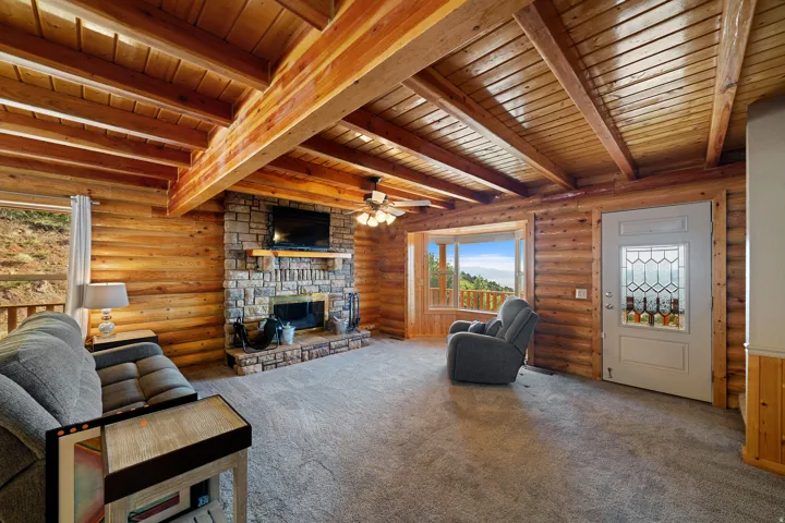 Spacious common area featuring log walls, exposed beam ceiling, stone-faced fireplace with wood mantel, large bay window, and light-colored entry door with decorative glass inserts