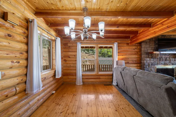 Living space featuring natural log walls, wood-finish flooring, exposed wood beams, and a stone fireplace with a wood mantle