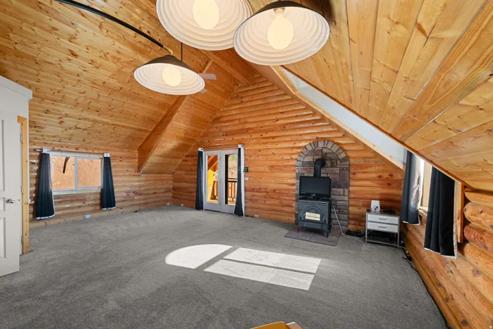 Expansive room featuring natural wood plank walls and ceilings, gray carpeting, a stone-accented wood-burning stove, and a pair of white French doors