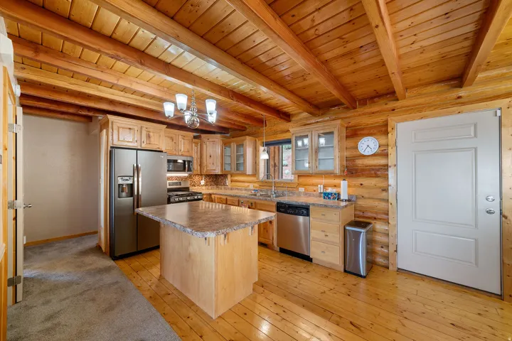Kitchen featuring wood-paneled walls and ceiling with exposed beams, wood-finish flooring, a central island with a stone-finish countertop, stainless steel appliances, and wood cabinetry with glass-front accents