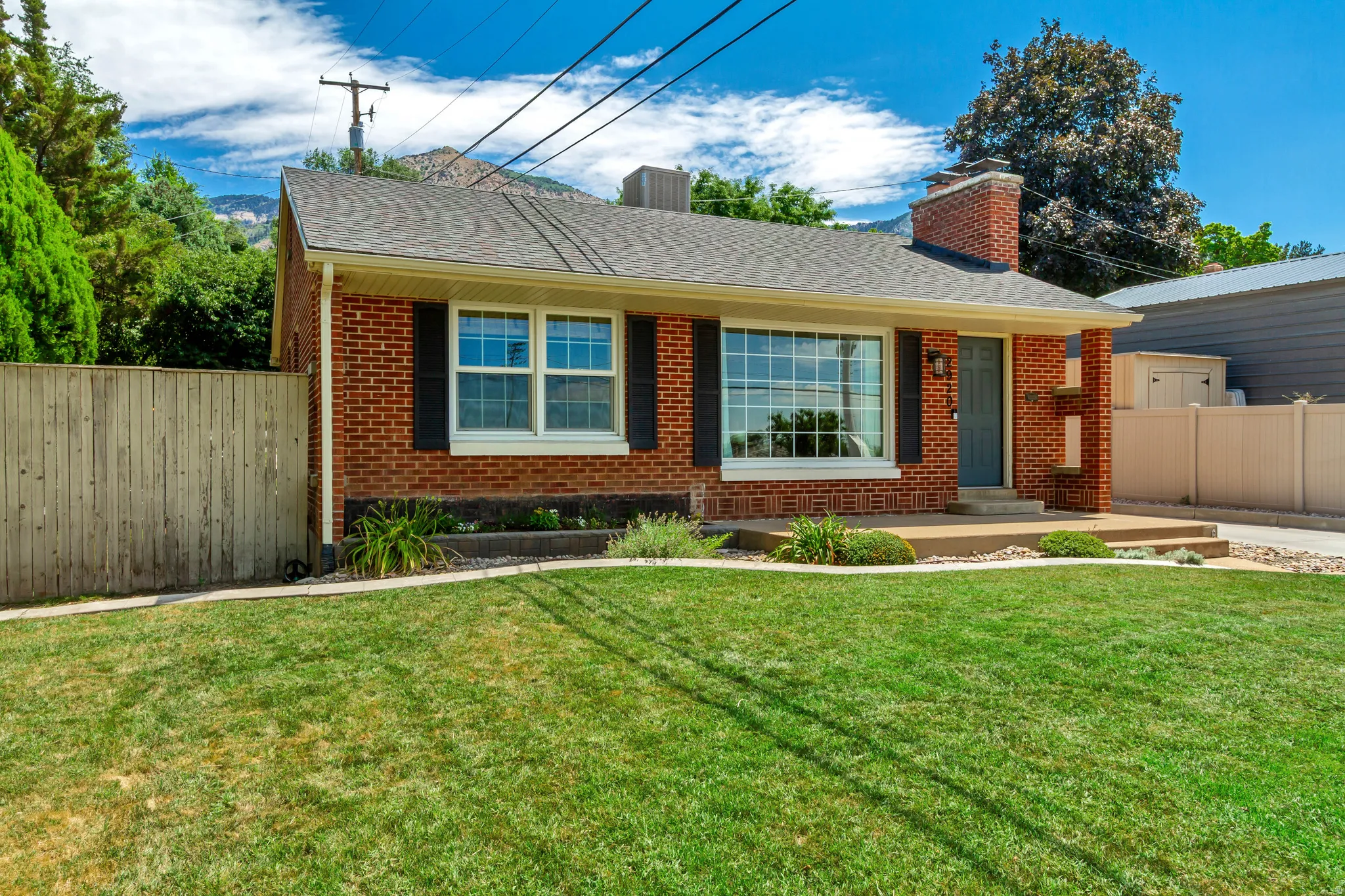Brick exterior home featuring a dark gray front door, a visible chimney, a large picture window with white framing, black shutters, and mature front landscaping