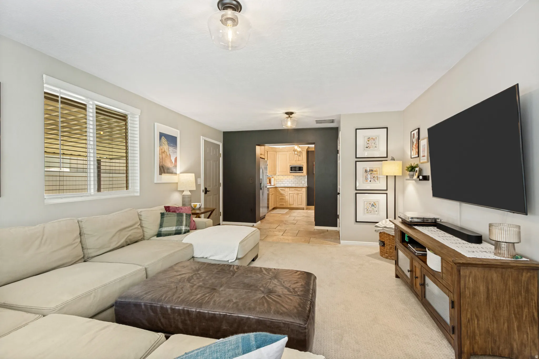 Carpeted living area featuring a large window with blinds, recessed lighting, and a doorway opening to a kitchen with tile flooring and wood cabinetry