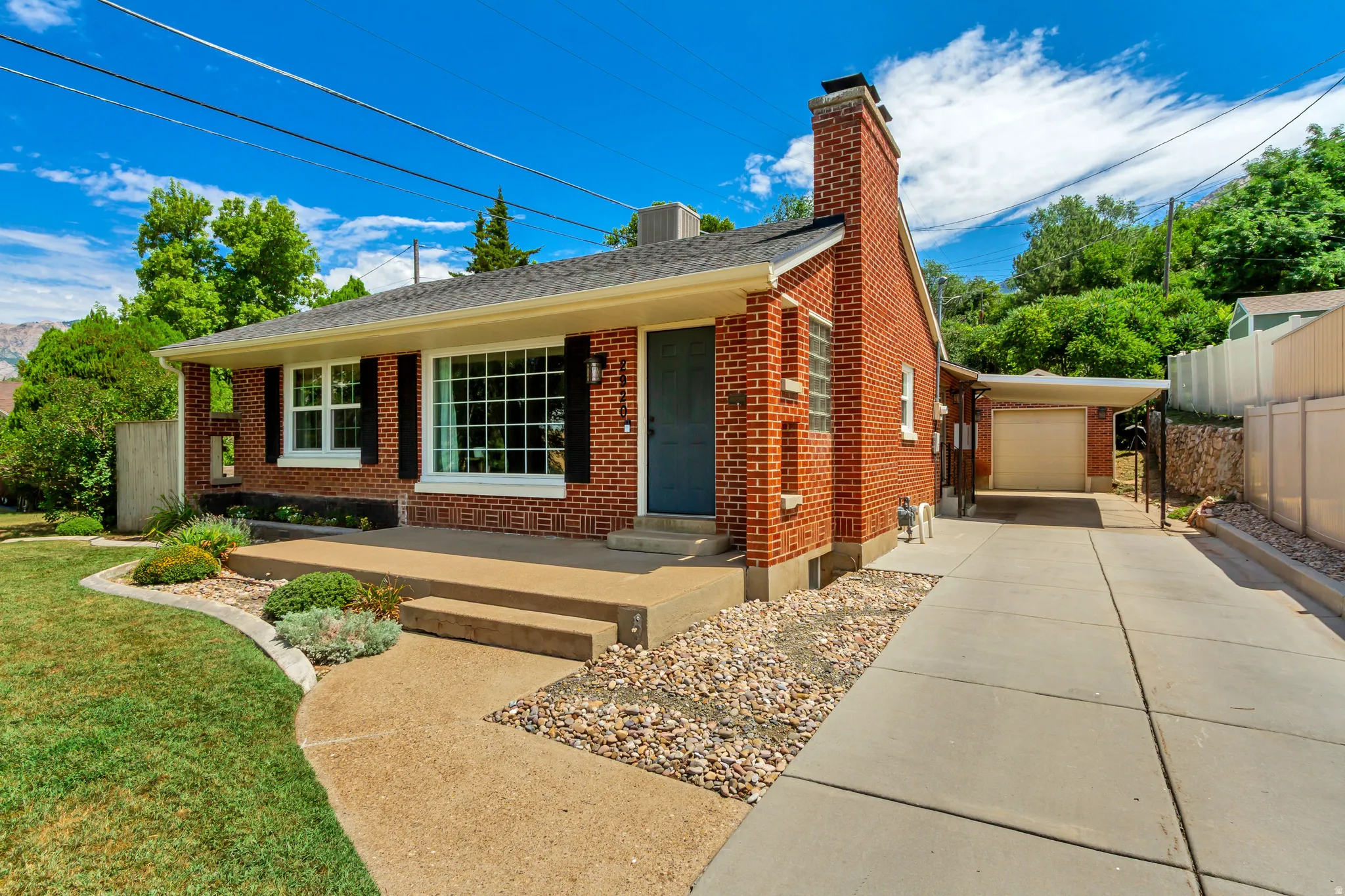 Traditional brick exterior with a prominent chimney, large picture window with black shutters, mature landscaping, and a concrete driveway leading to a covered carport and attached garage