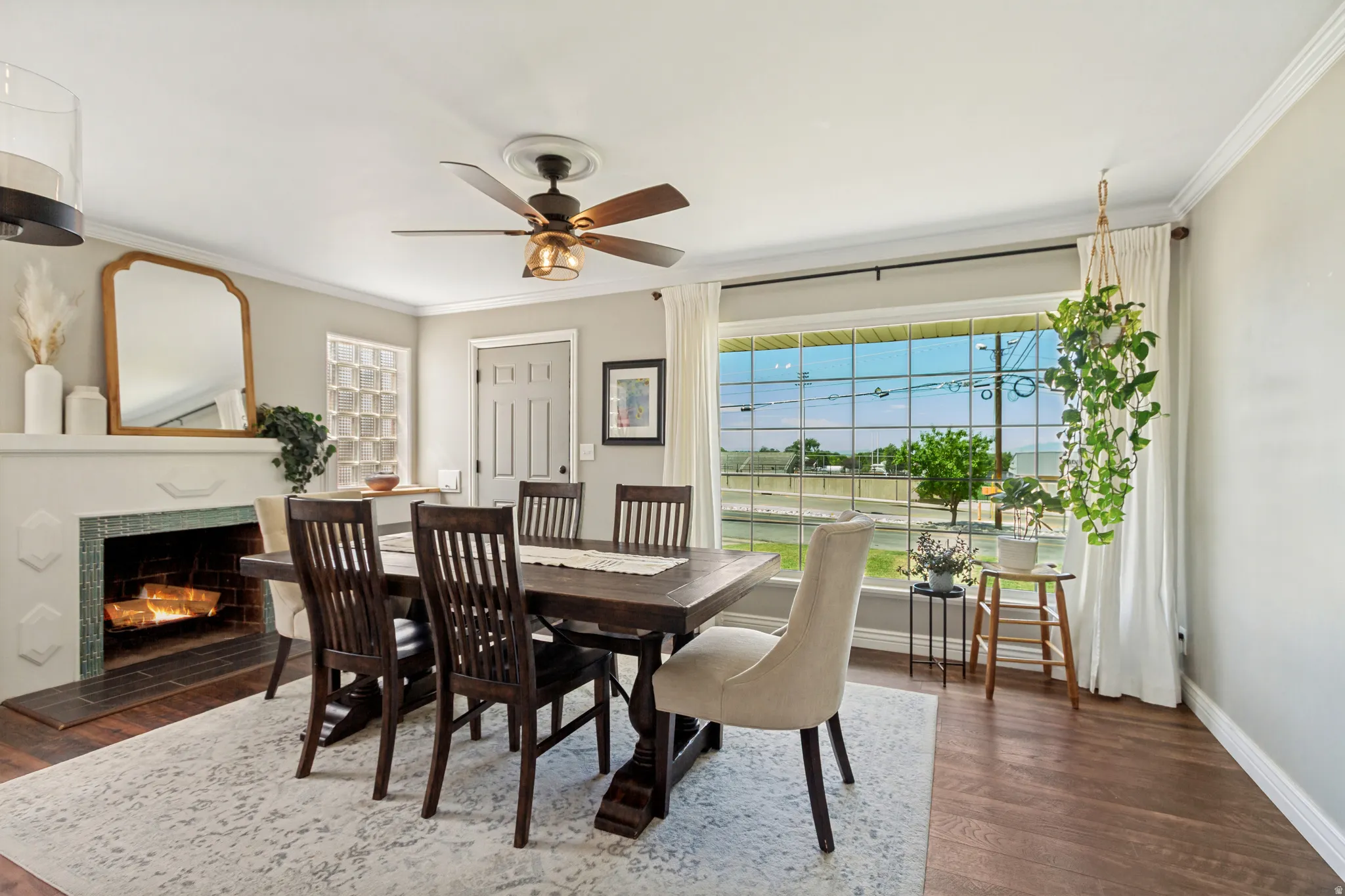 Dining space featuring wood-finish flooring, a decorative fireplace with tile surround, a ceiling fan with light fixture, and a large window with white curtain panels