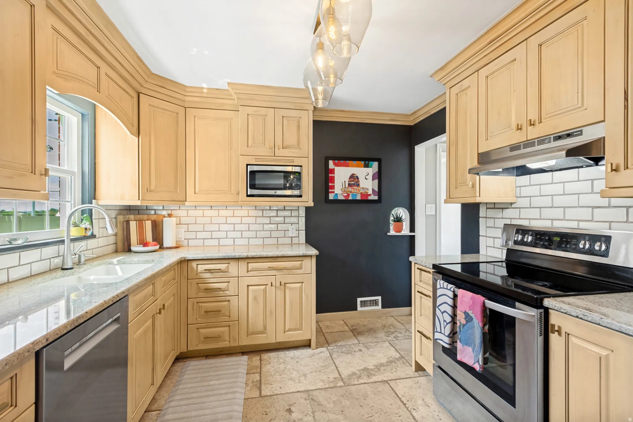 Light-toned cabinetry with crown molding, granite countertops, subway tile backsplash, stainless steel appliances, and large format stone flooring