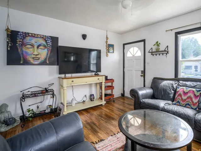Spacious living area featuring warm wood-finish flooring, a white paneled entry door with an arched window, and a bright window with dark trim