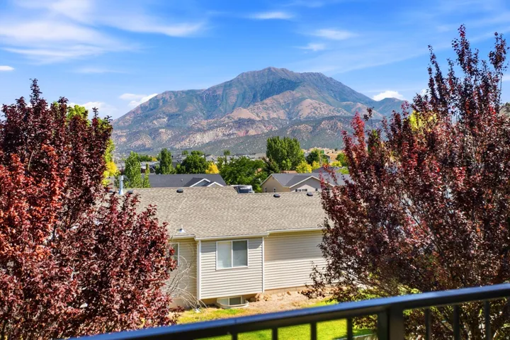 Mountain backdrop framing residential properties under a clear sky, featuring a gray railing, red-leafed trees, and a light-colored siding structure with a shingled roof