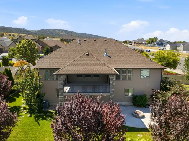 Rear exterior featuring a covered balcony with stone pillars, a lower-level patio with a fire pit, and a pitched roof with asphalt shingles