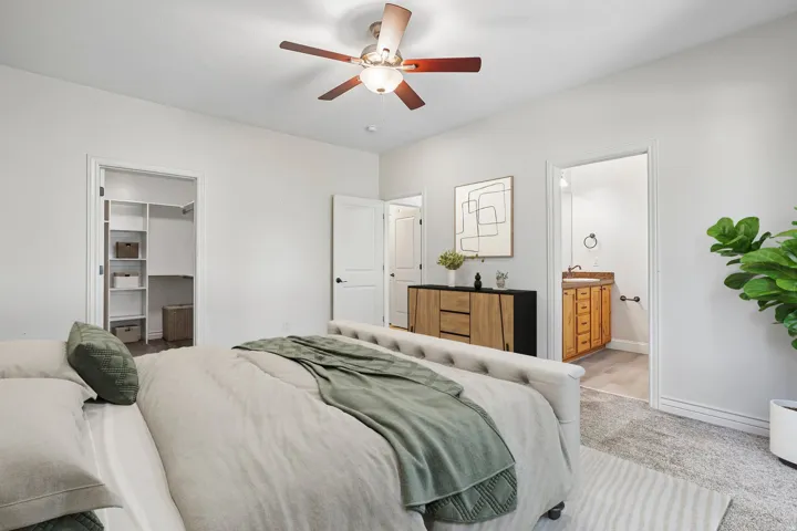 Carpeted room featuring a ceiling fan with integrated lighting, a walk-in closet with built-in shelving, an entryway to a bathroom with a wood-finish vanity, and white baseboards
