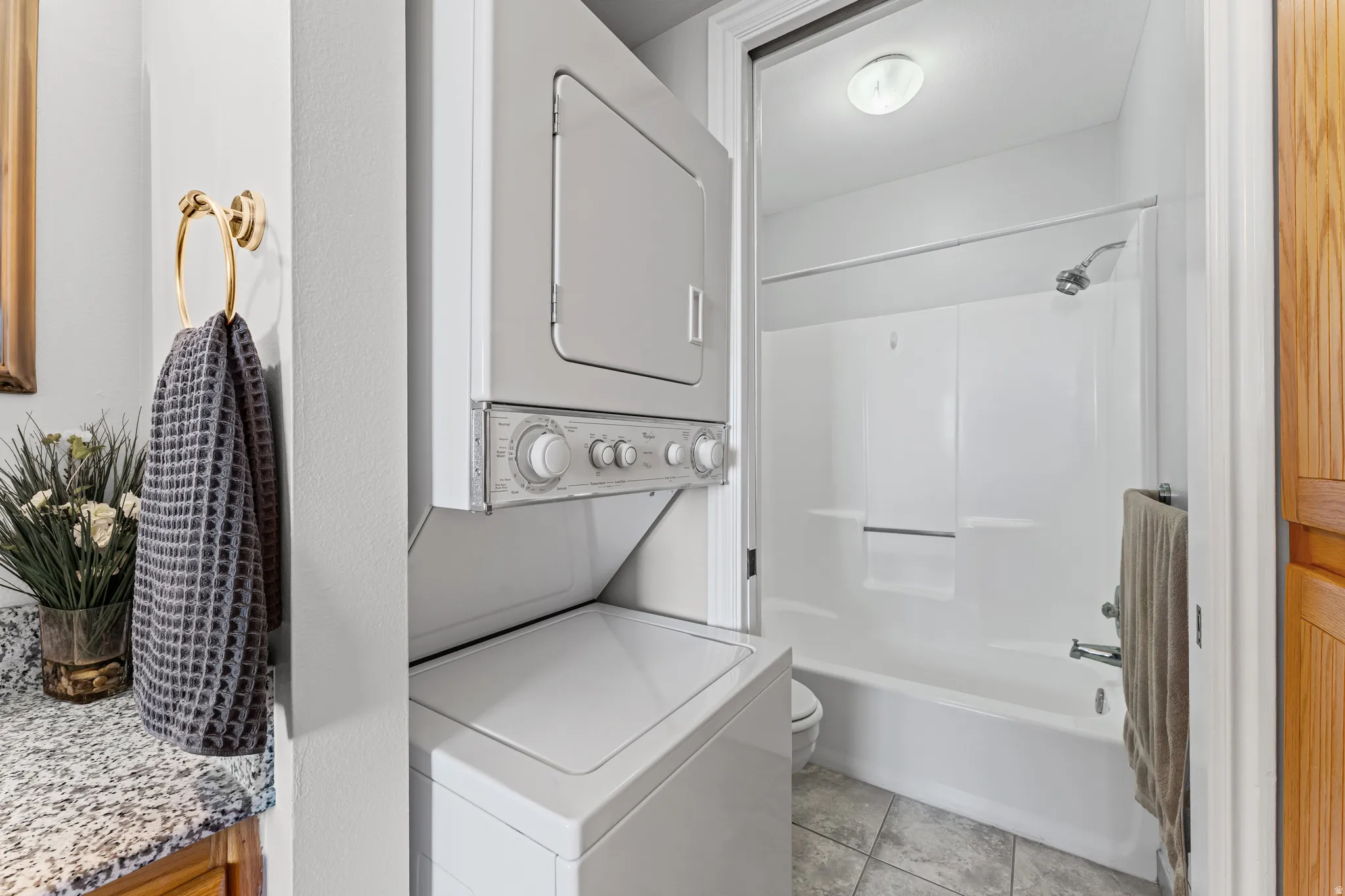 Compact laundry area with stackable washer and dryer, adjacent to a full bathroom featuring a white tub/shower combination, tile flooring, and a ceiling-mounted light fixture