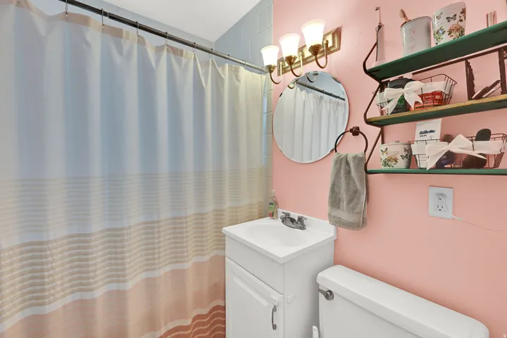 Charming bathroom featuring soft pink walls, a white pedestal sink with integrated storage, and a round mirror with brass-finished lighting fixture