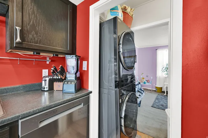 Convenient laundry room featuring stacked washer and dryer units, vibrant red accent walls, dark countertop workspace with integrated appliances, and direct access to adjacent living spaces