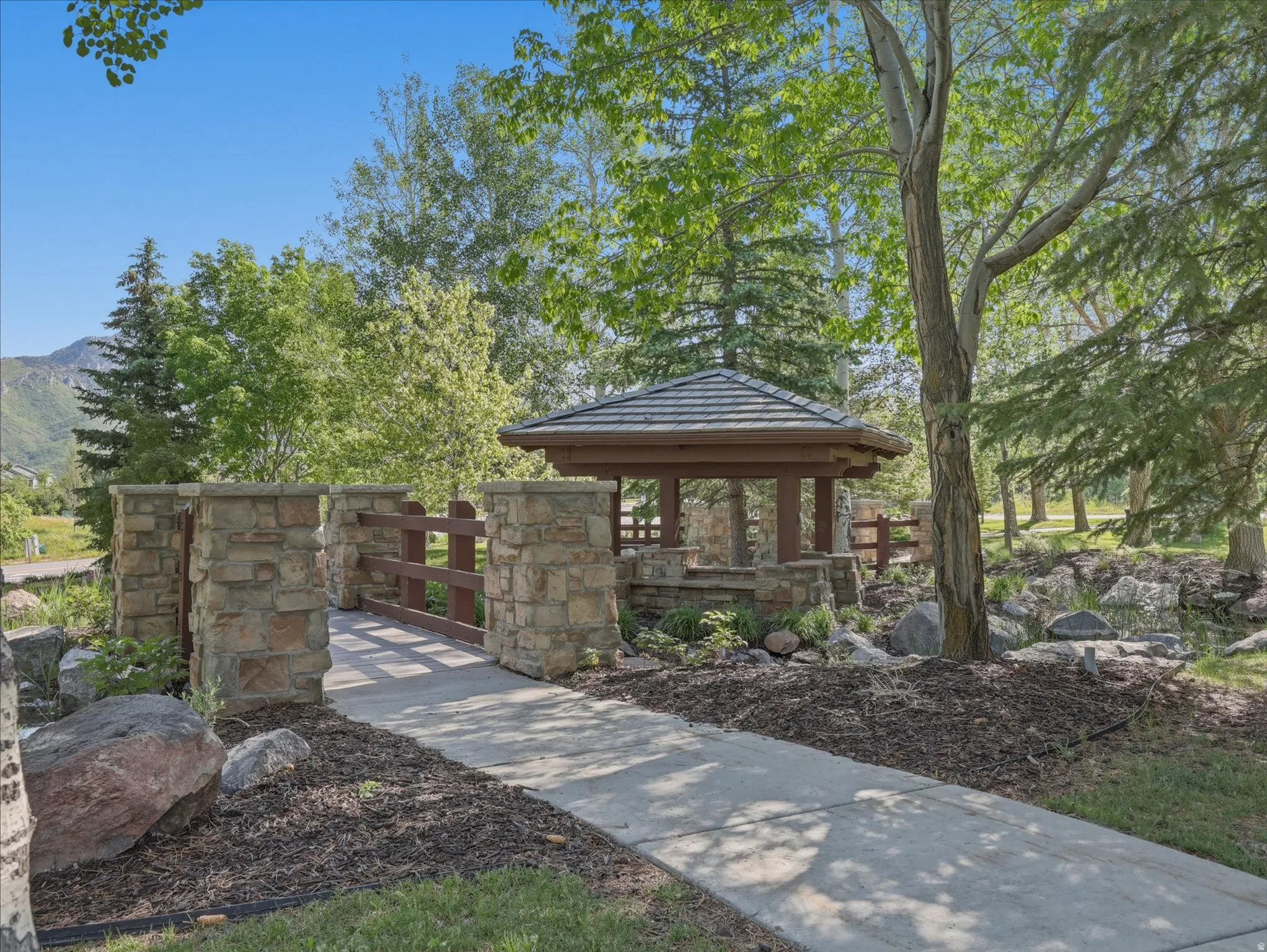 Landscaped outdoor area featuring a wood gazebo with a shingled roof, stone pillars, a wooden bridge, and a paved walkway
