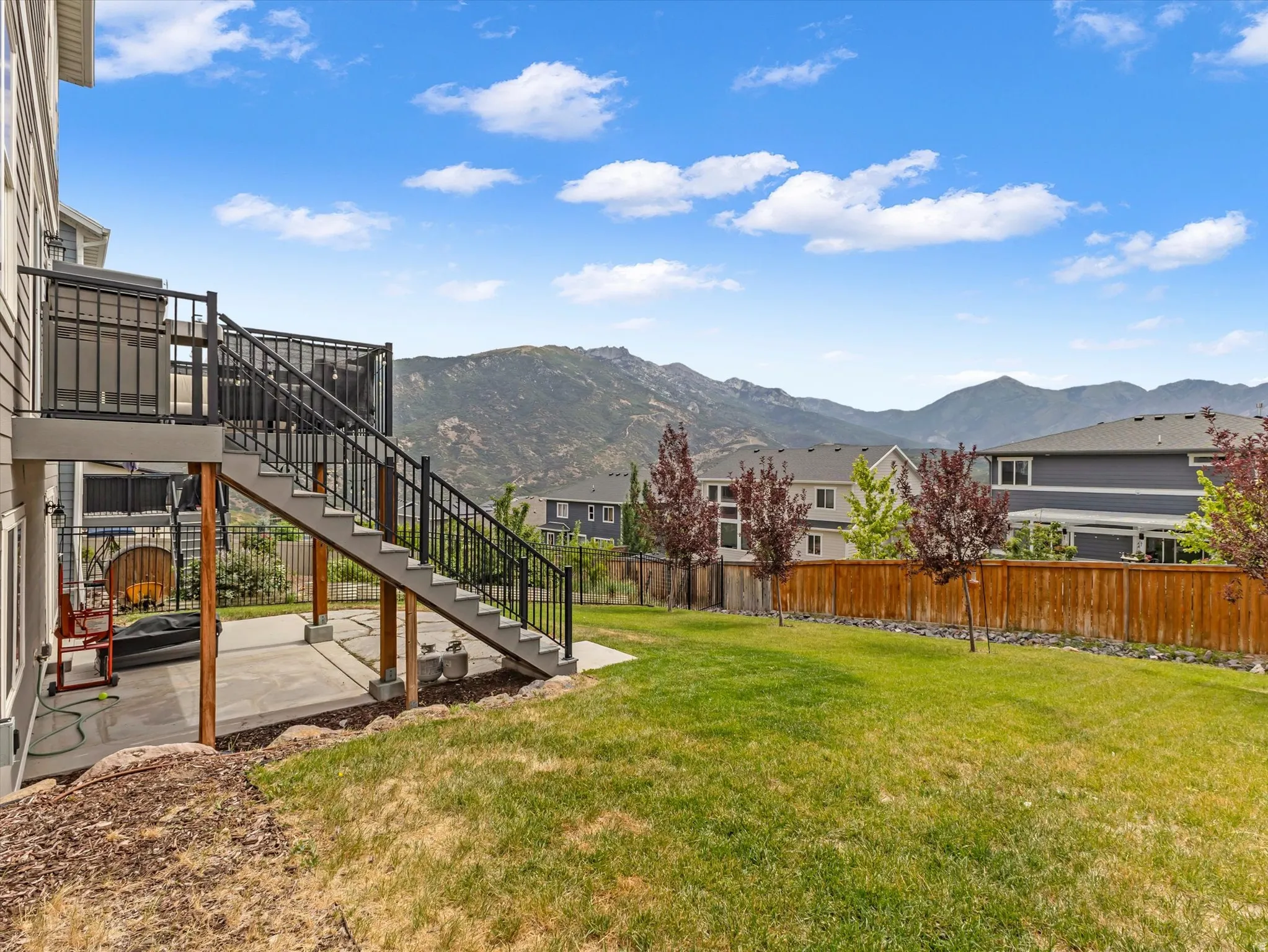 Spacious backyard featuring a multi-level deck, concrete patio, mature trees, wood fence, and mountain backdrop