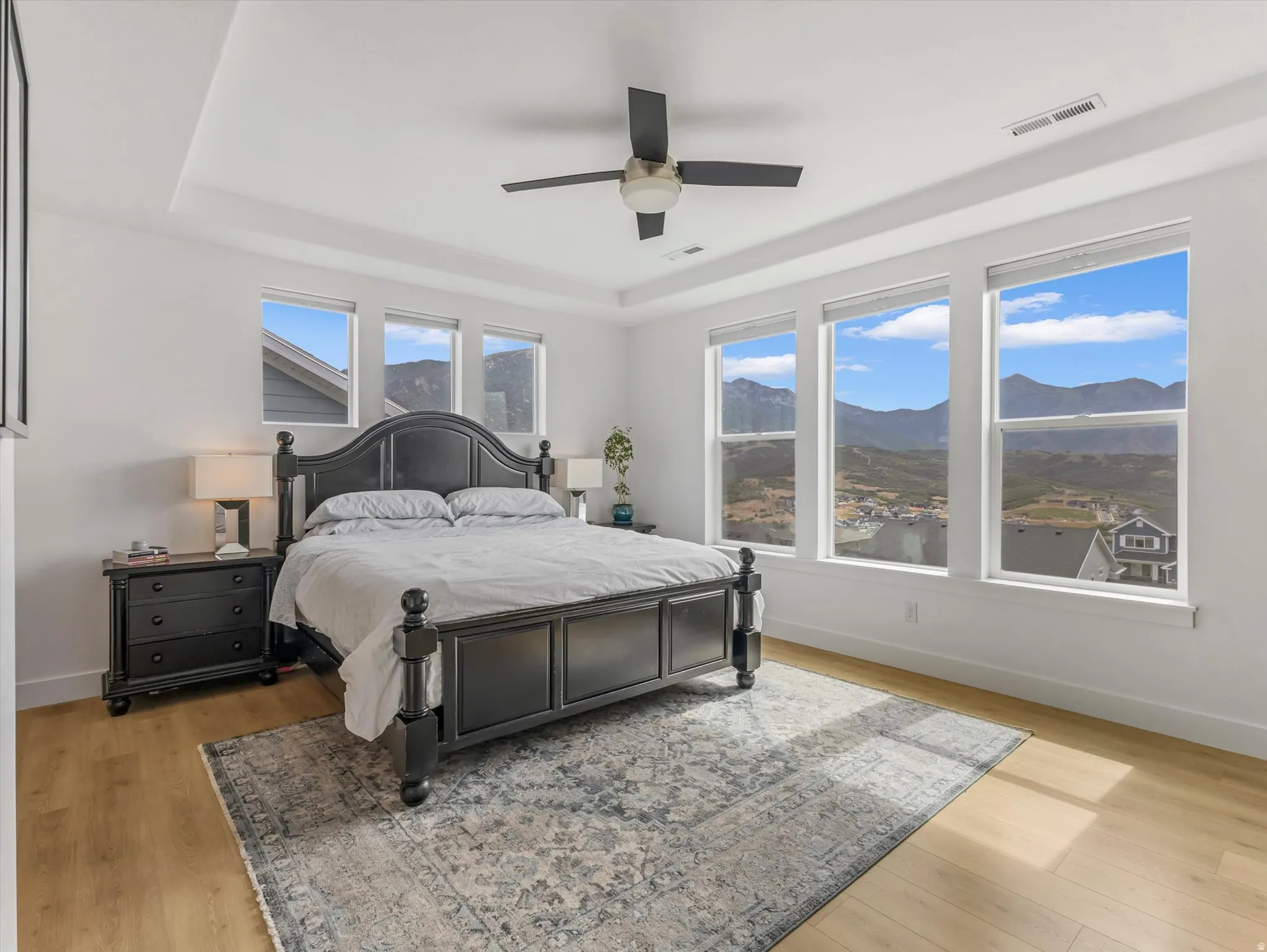 Spacious room featuring multiple large windows, light wood-finish flooring, a contemporary ceiling fan, and a tray ceiling detail