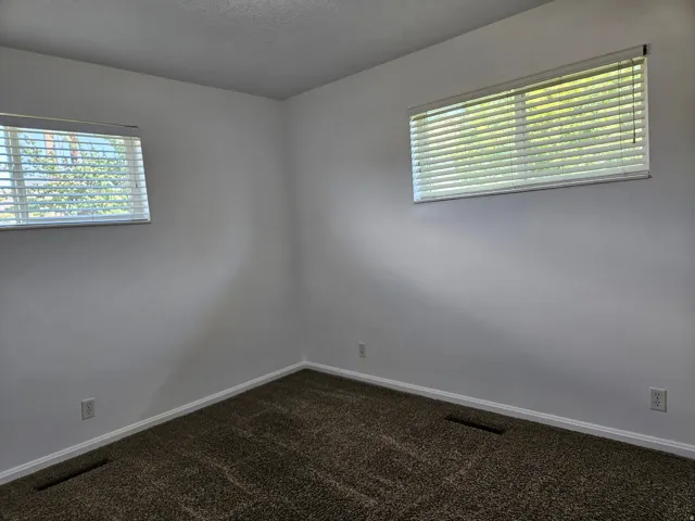 Spacious room featuring neutral wall tones, two windows with horizontal blinds, textured carpeting, and white baseboards