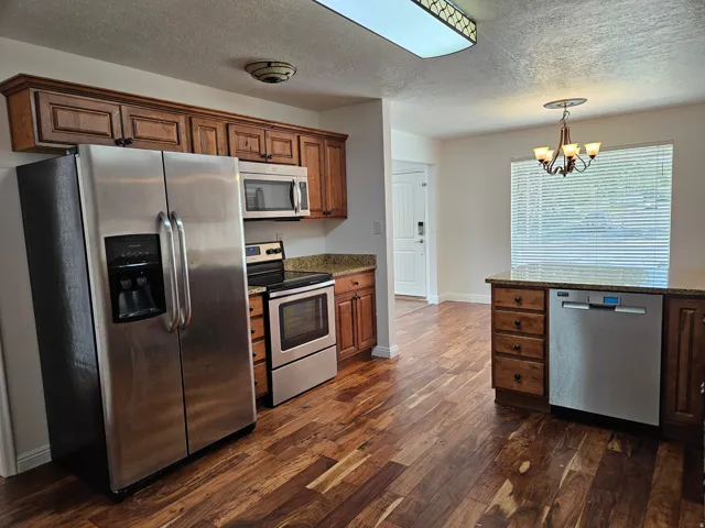 Kitchen featuring wood-finish flooring, stainless steel appliances, wood cabinetry, stone countertops, and a dining area with a chandelier
