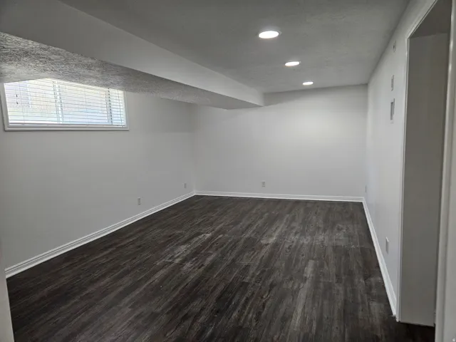 Spacious room featuring dark wood-finish flooring, recessed lighting, a window with blinds, white baseboards, and freshly painted white walls