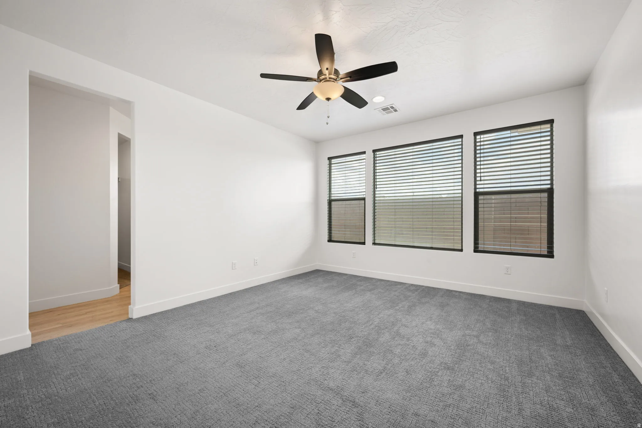 Spacious room featuring textured gray carpeting, multiple windows with blinds, a ceiling fan with integrated lighting, and crisp white baseboards