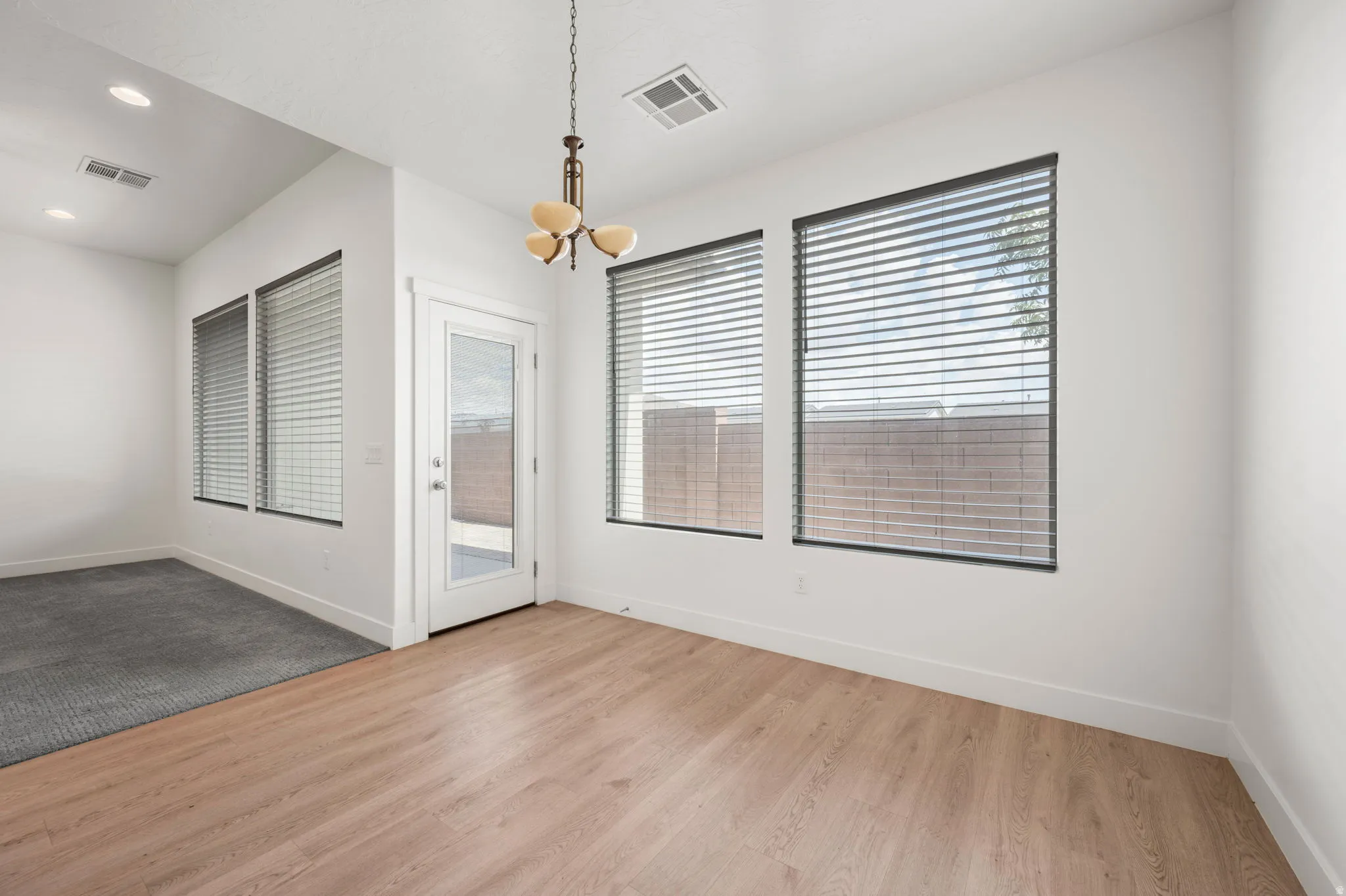 Spacious area featuring light wood-finish flooring, multiple windows with blinds, a glass-paned exterior door, and a decorative chandelier