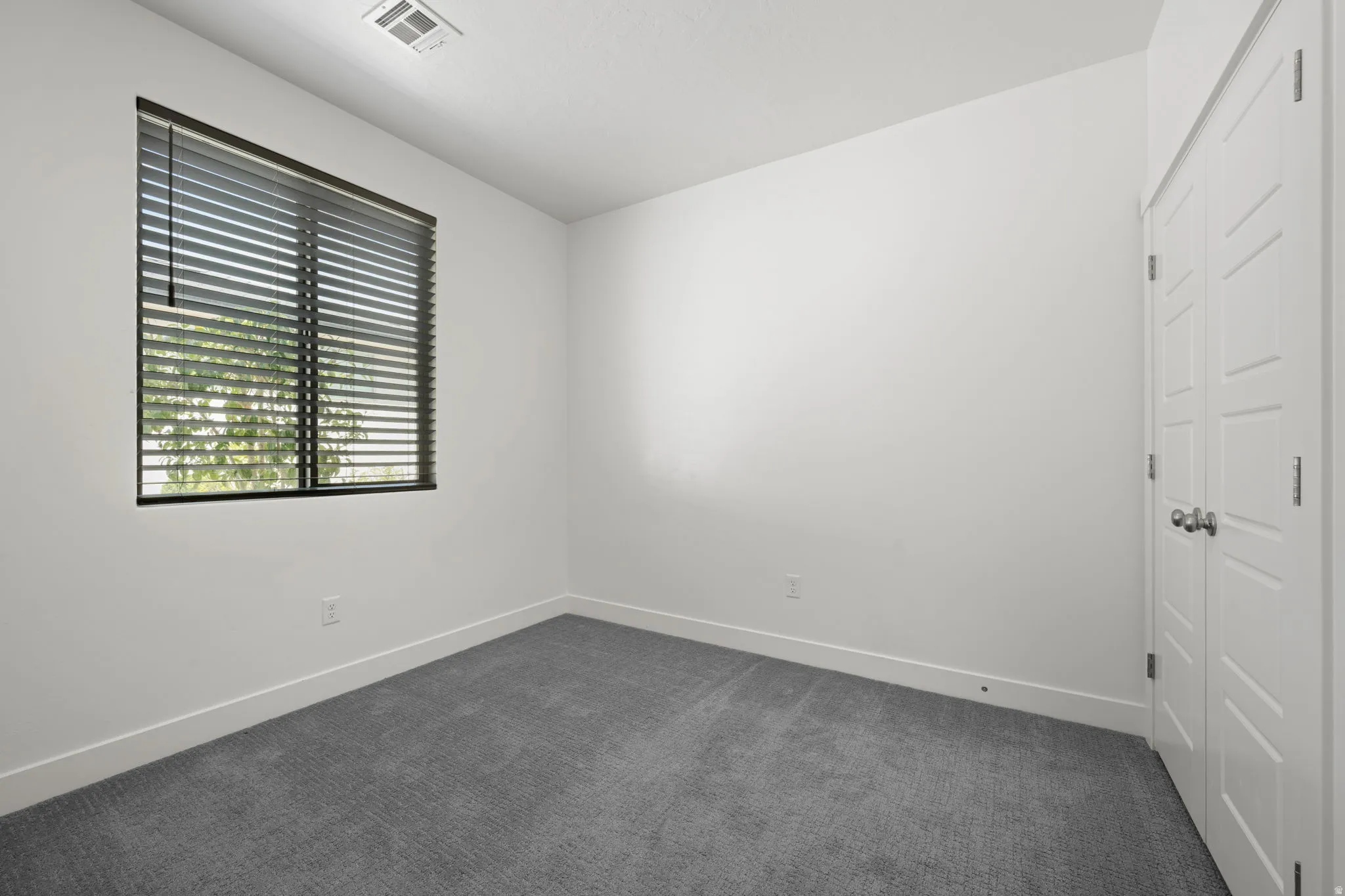 Neutral color palette with white walls, grey carpet, a window with horizontal blinds, white baseboards, and a white paneled closet door with silver hardware