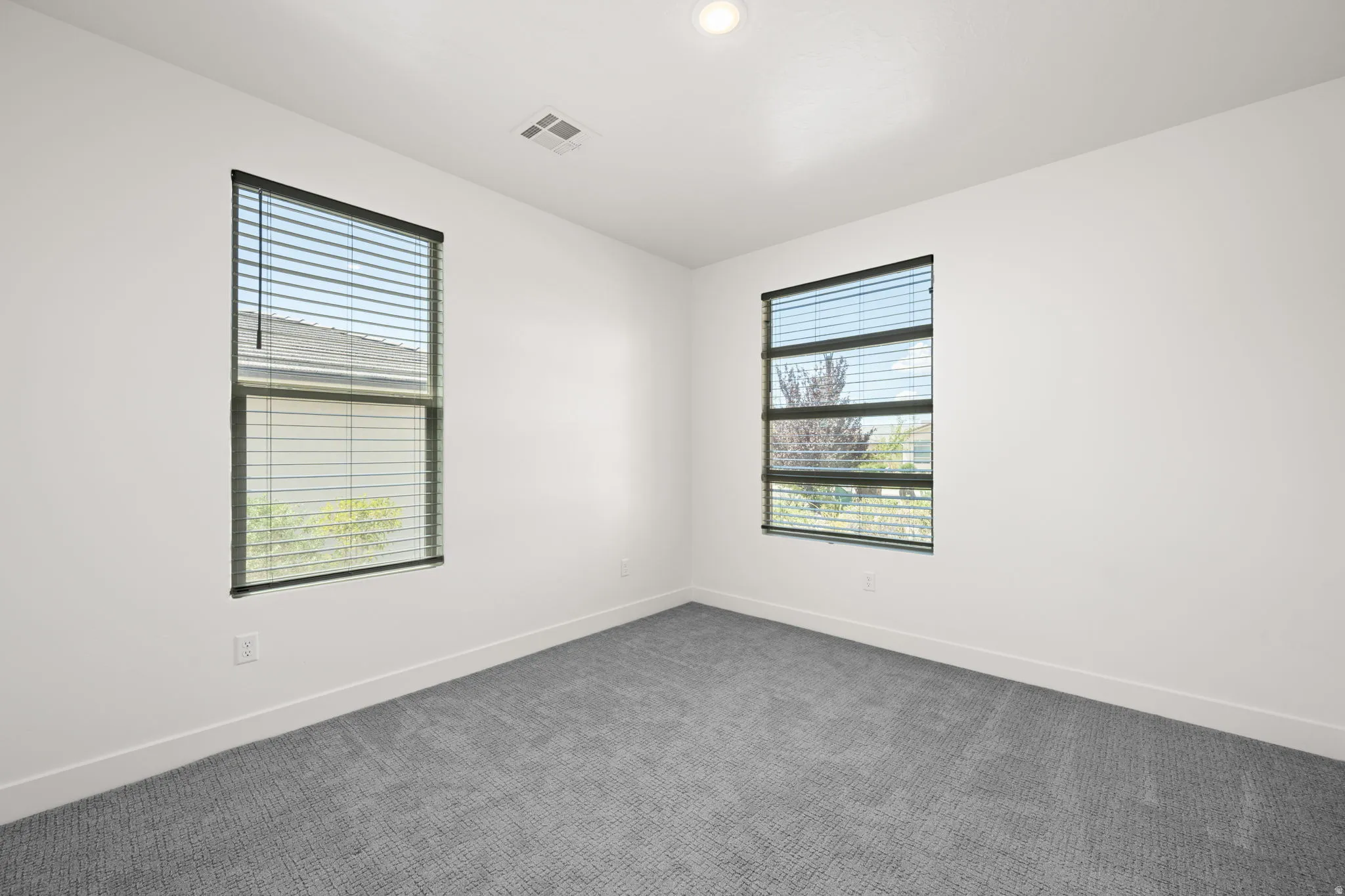 Bright room featuring two windows with horizontal blinds, neutral gray carpeting, white baseboards, and a recessed ceiling light fixture