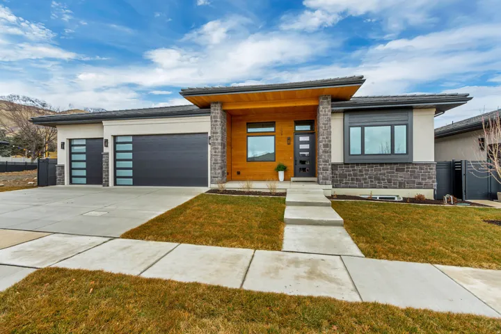 Modern ranch-style exterior featuring a multi-textured facade, stone accents, cedar-finish siding, a broad concrete driveway, and an attached three-car garage with contemporary glass-paneled doors