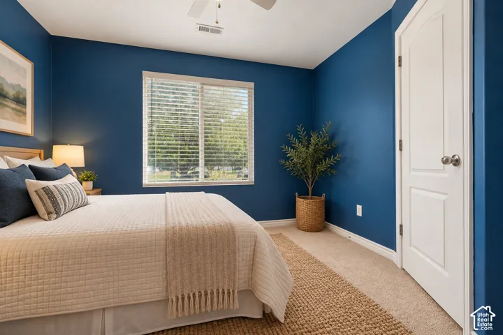 1st Bedroom featuring neutral carpeting, sliding window with horizontal blinds, ceiling fan, and a solid-core interior door with metallic hardware. Virtually Staged.