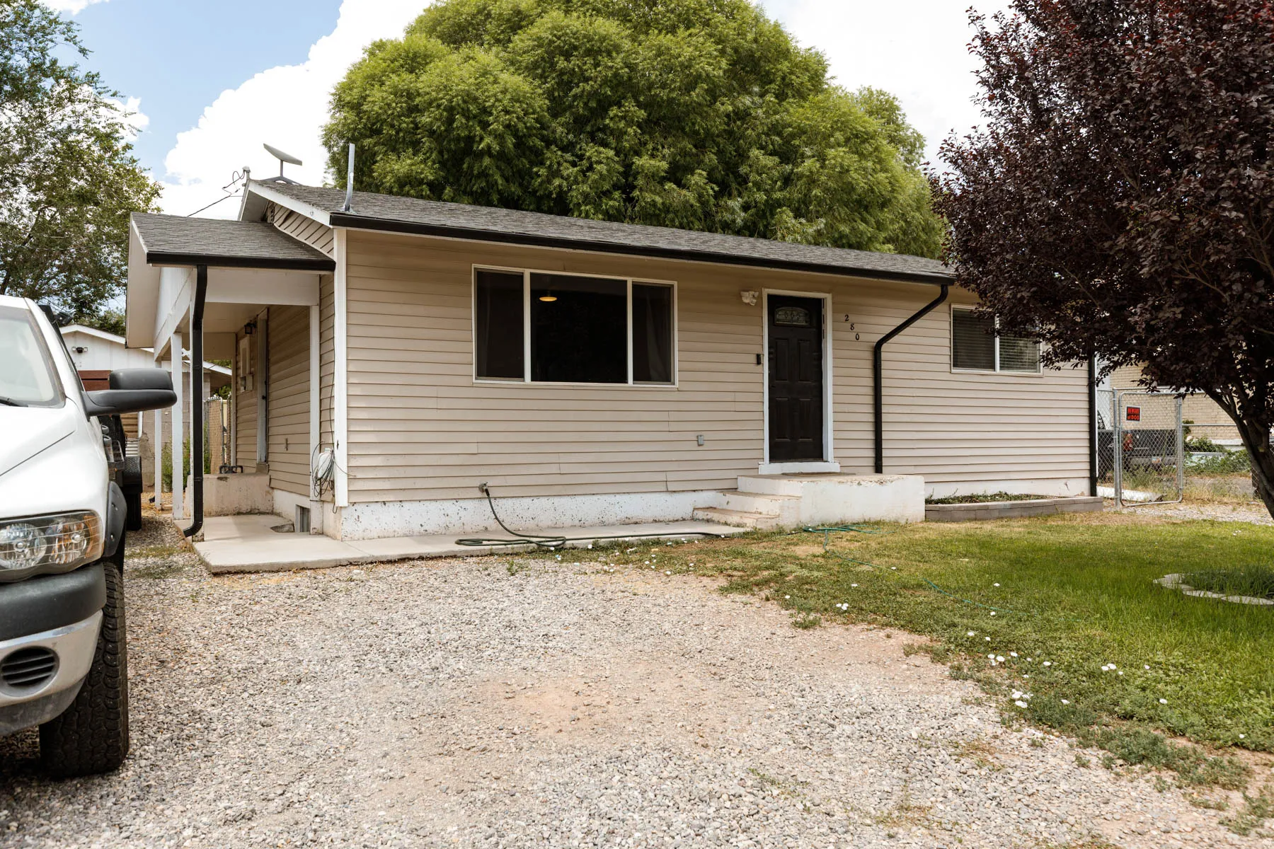 Single-story property featuring tan horizontal siding, a black front door, a gravel driveway, and a partially grassed front yard