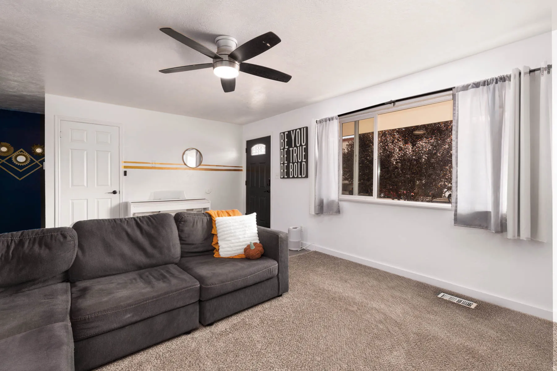 Living area featuring a ceiling fan with integrated lighting, a large window with sheers and drapes, a paneled door, and neutral-toned carpeting