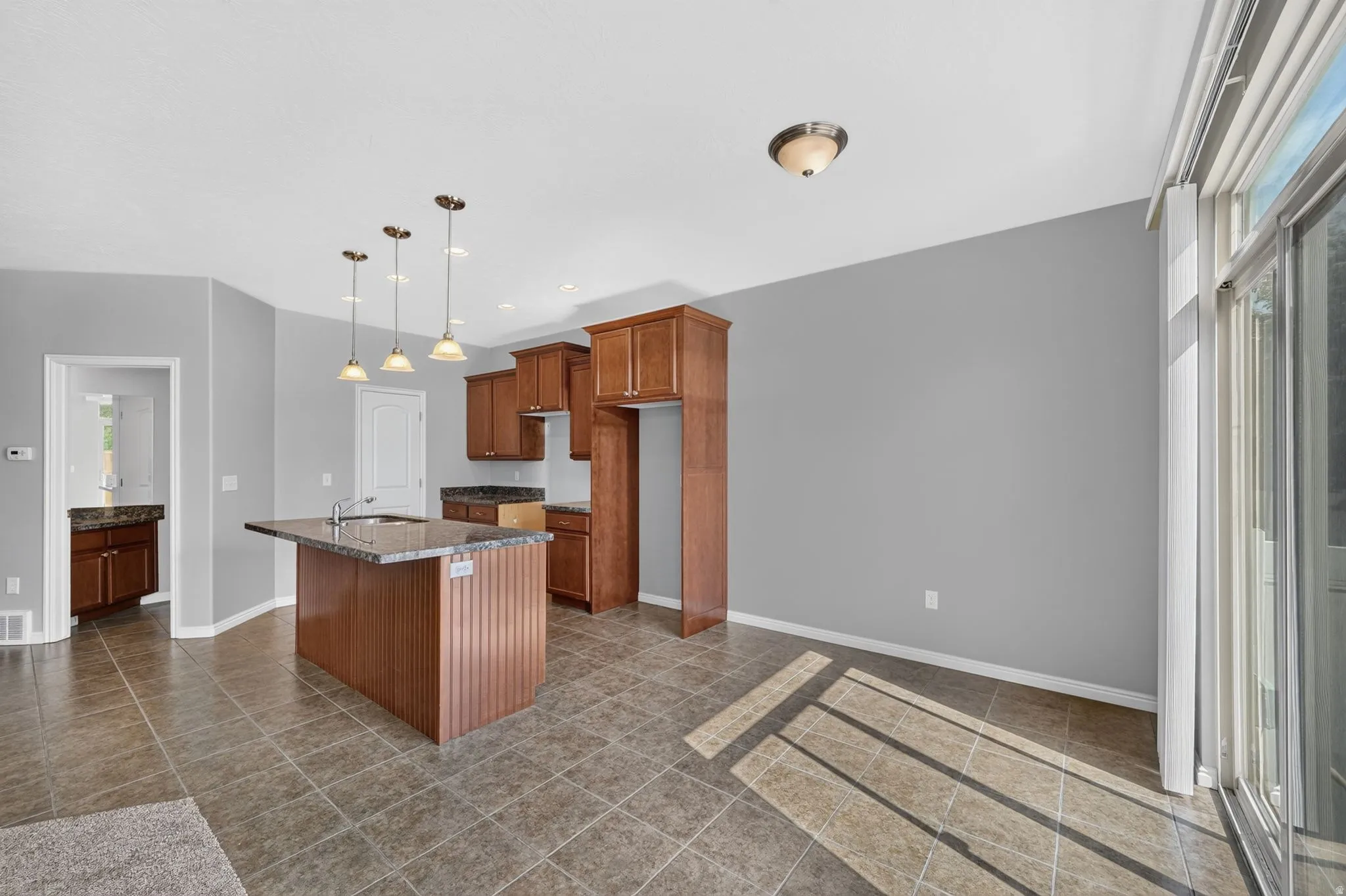 Kitchen featuring an island with a built-in sink, pendant lighting, wood cabinetry, stone countertops, and tile flooring