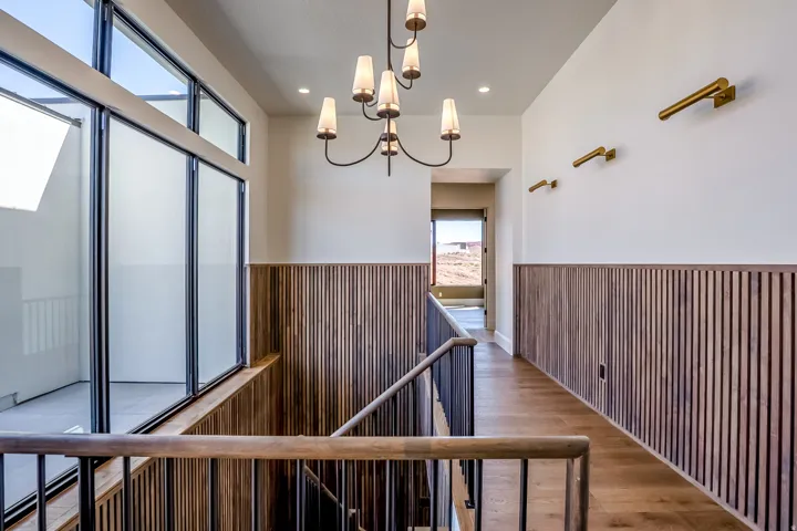 Wood-finish flooring, staircase with metal and wood-finish railing, wood-slat wall paneling, modern chandelier, and large windows with transom panels