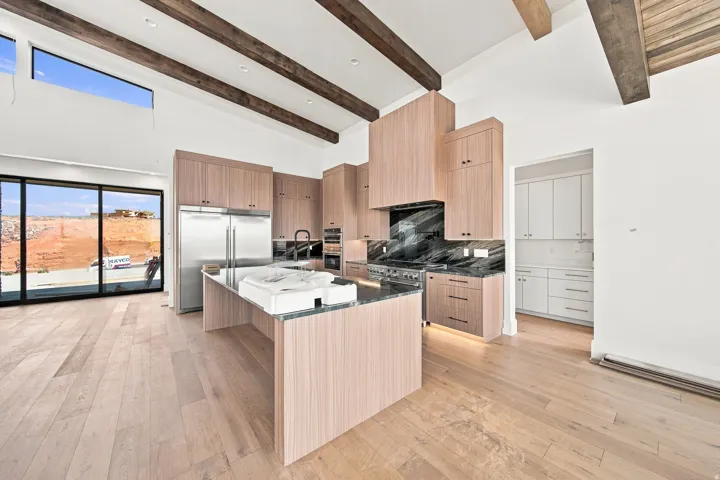 Vaulted ceiling with exposed wood beams, wood-finish flooring, full-height sliding glass doors, kitchen island with dark stone countertop, and built-in stainless steel appliances